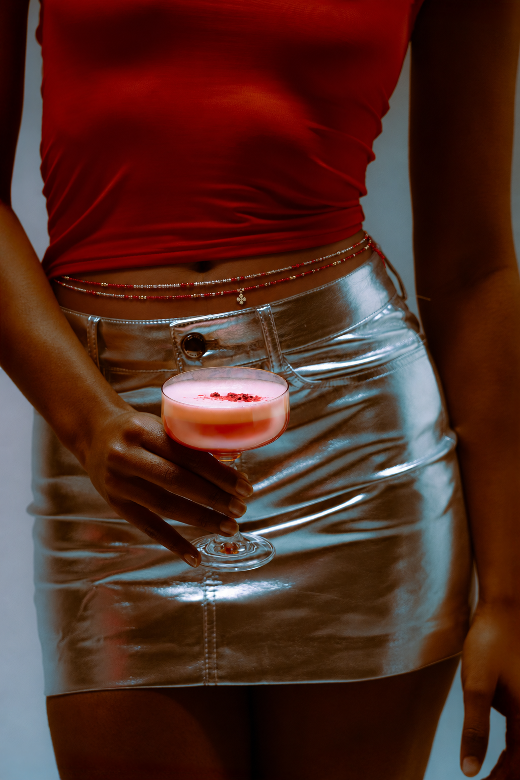 Donkergekleurde vrouw in rode top en zilveren rok met rode en zilveren waistbeads rond haar middel, terwijl ze een roze cocktail vasthoudt.